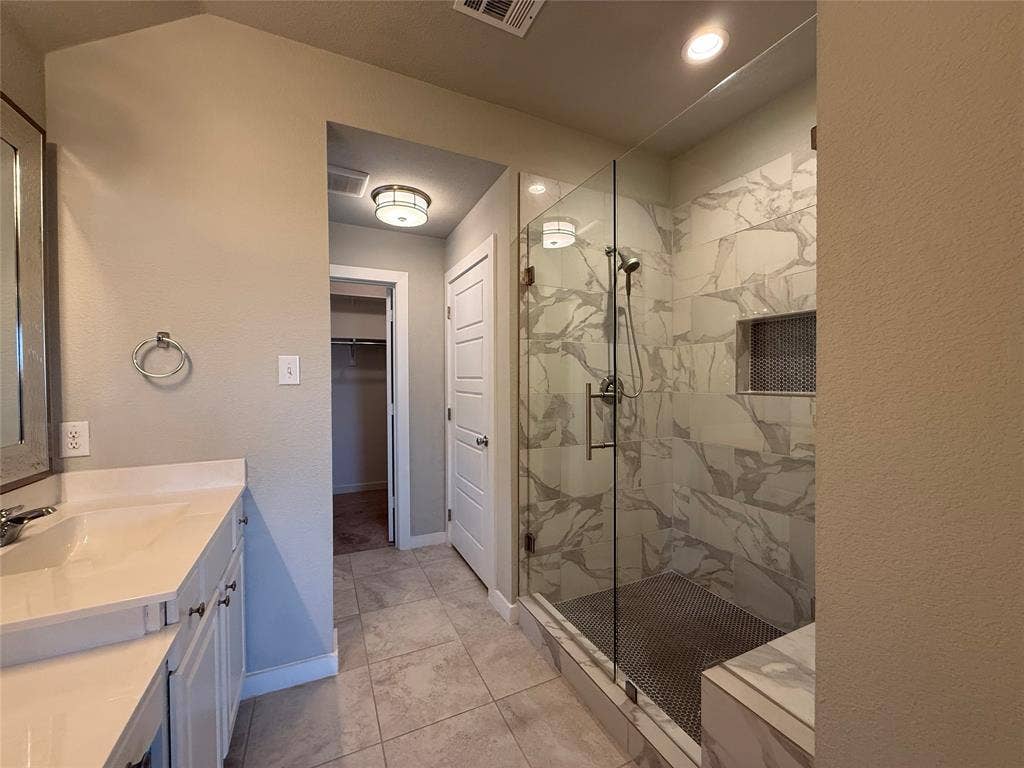 1702 Sagebrush Trail, Wylie, TX 75098 Walk-in shower featuring large-format tile surround, glass enclosure, built-in recessed niche, and stone-finish floor tiling, complemented by a white vanity with integrated basin