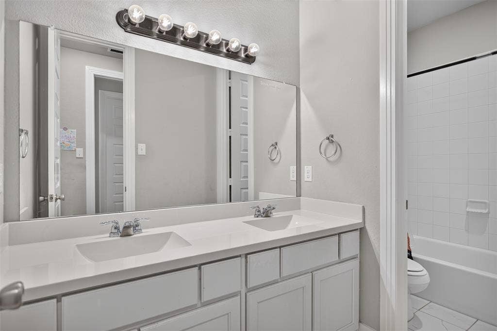 1103 Walford Drive, Forney, TX 75126 Double vanity featuring solid-surface countertops, dual integrated sinks, an expansive wall-to-wall mirror, and overhead multi-light fixture