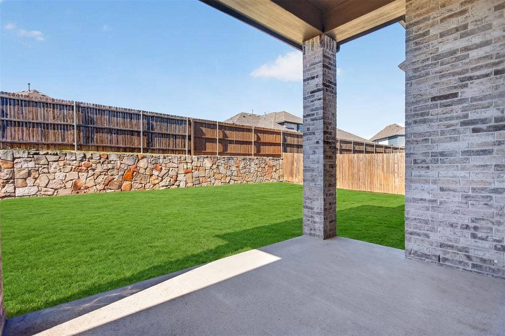 1103 Walford Drive, Forney, TX 75126 Covered patio featuring brick pillar supports, concrete flooring, manicured lawn, stone retaining wall, and wood fencing
