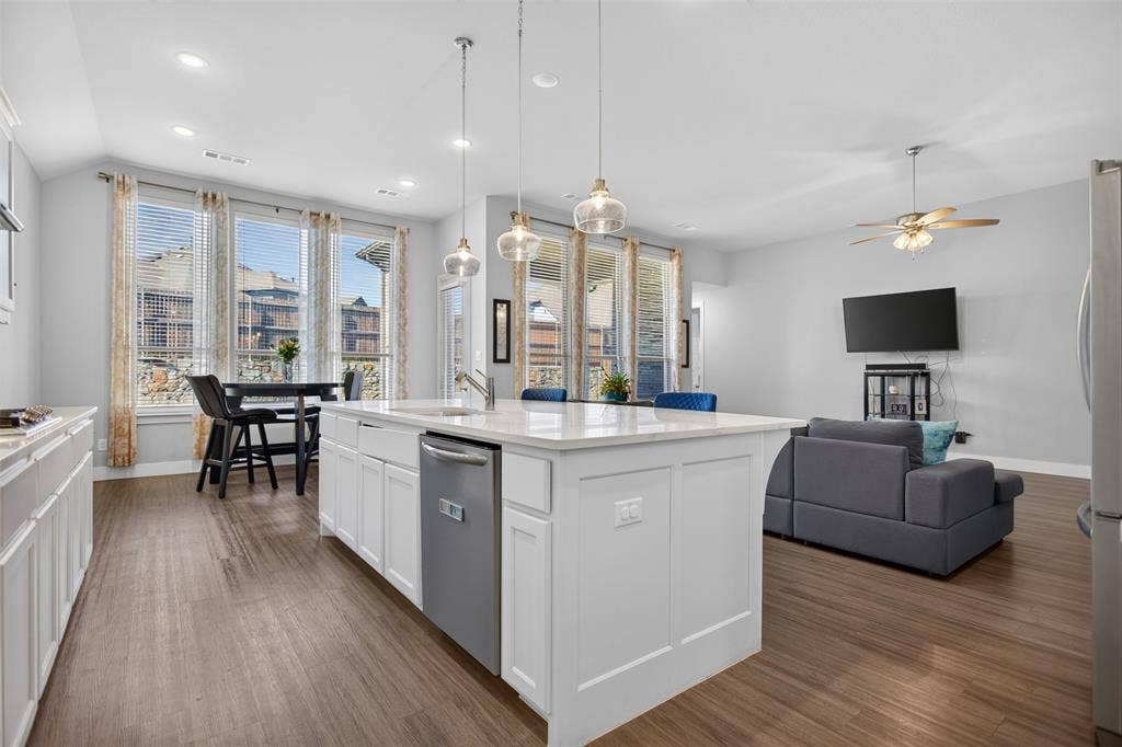 1103 Walford Drive, Forney, TX 75126 Kitchen featuring a large central island with white cabinetry, light-toned stone-finish countertop, stainless steel dishwasher, and three glass pendant lights, complemented by wood-finish flooring and multiple tall windows