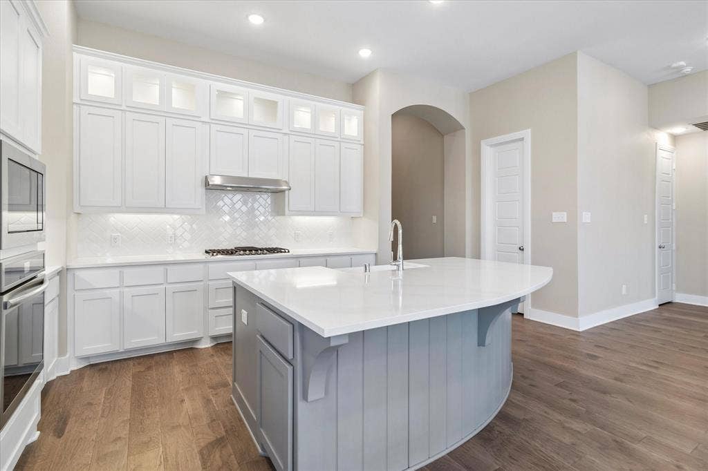 Kitchen featuring white shaker-style cabinetry with stacked uppers, stone-surface countertops, a curved center island with beadboard detailing, wood-finish flooring, and a stainless steel built-in oven and microwave