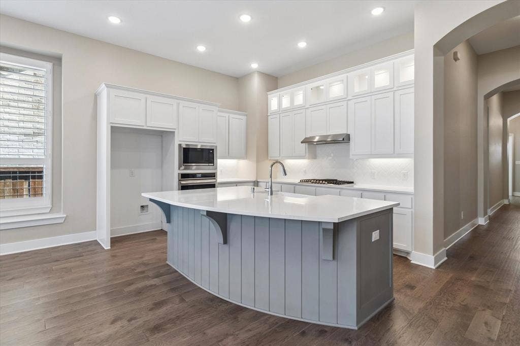 Kitchen featuring white cabinetry with glass-front uppers, a contrasting gray-paneled island with a stone-finish countertop, recessed lighting, and wood-finish flooring