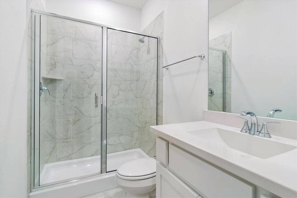 Bathroom featuring a walk-in shower with glass-enclosure doors, light-toned stone-finish tile surround, built-in shelving, white cabinetry with a solid-surface countertop, and a wall-mounted mirror