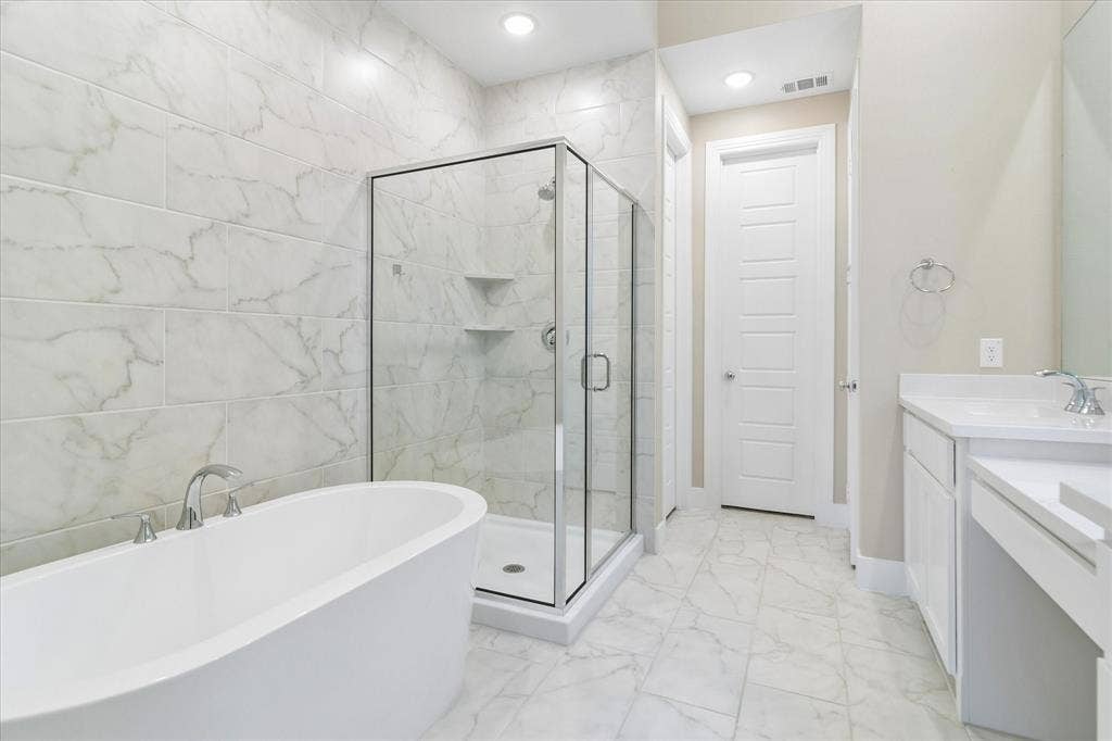 Freestanding white soaking tub, glass-enclosed shower with full-height stone-finish wall tile, white vanity cabinetry with surface-mounted sink, and stone-finish floor tiling