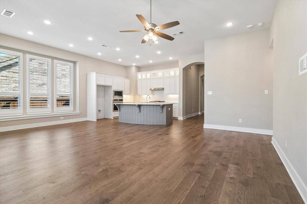 Open-concept interior featuring wood-finish flooring, recessed lighting, a center island with a stone countertop, white shaker-style cabinetry, and large windows with plantation shutters