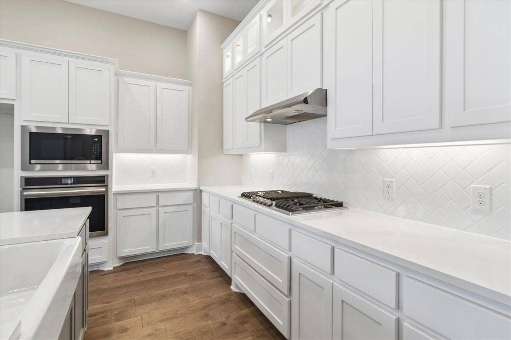 Kitchen featuring white shaker-style cabinetry with glass-front uppers, solid surface countertops, a herringbone tile backsplash, integrated stainless steel appliances, and wood-finish flooring