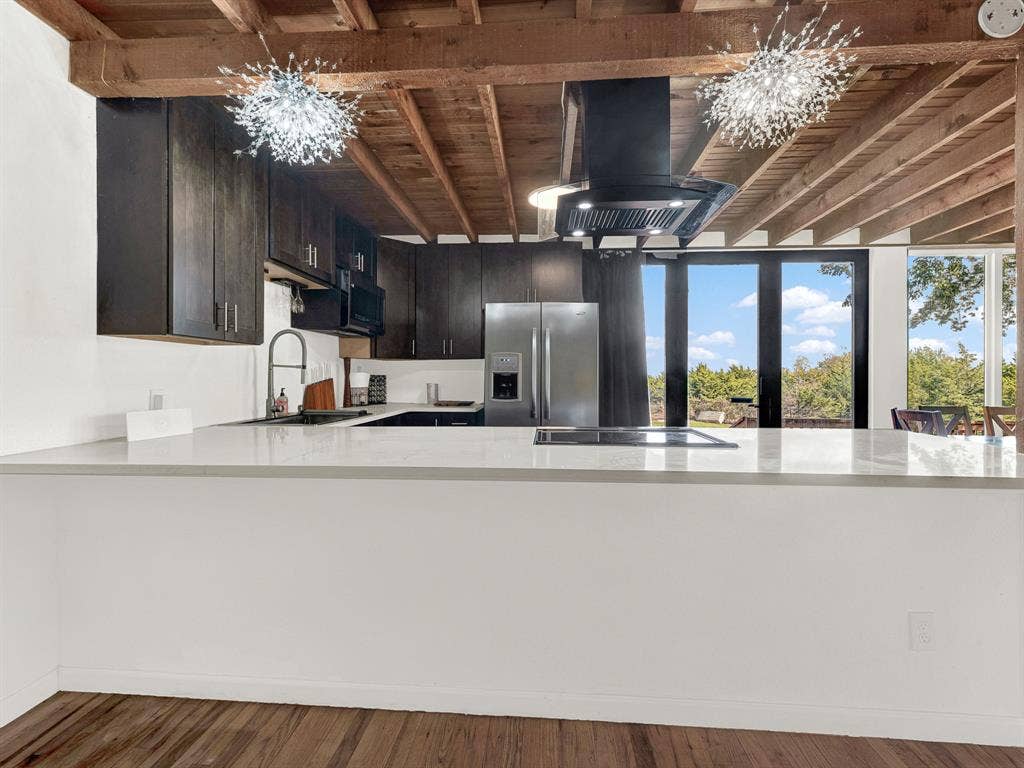 Kitchen featuring dark wood cabinetry, expansive stone-surface peninsula, exposed wood ceiling beams, stainless steel appliances, and multi-paned glass exterior doors