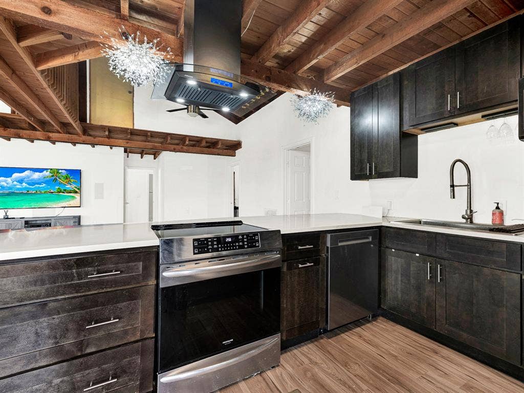 Kitchen featuring dark wood cabinetry, solid surface countertops, stainless steel appliances, a center-mounted range hood, and an exposed beam vaulted ceiling