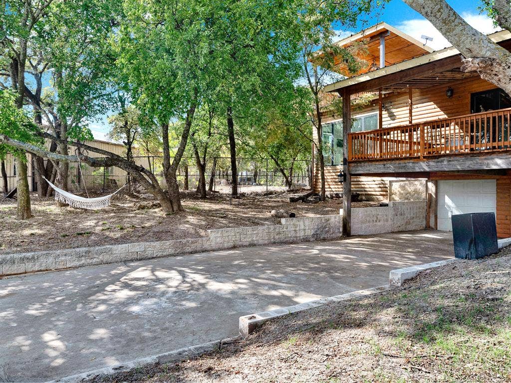 Wood-sided exterior featuring an elevated wood deck, concrete driveway, stone-block retaining wall, and mature trees