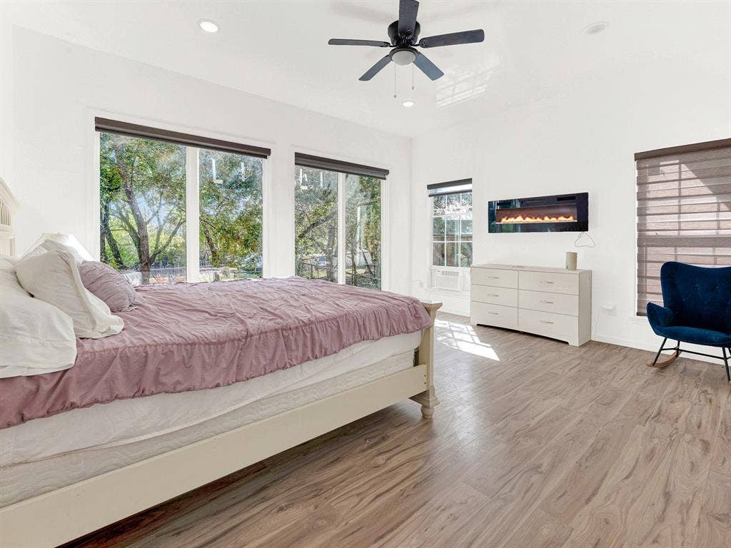 Wood-finish flooring, wall-mounted electric fireplace, ceiling fan, and large windows with integrated shades