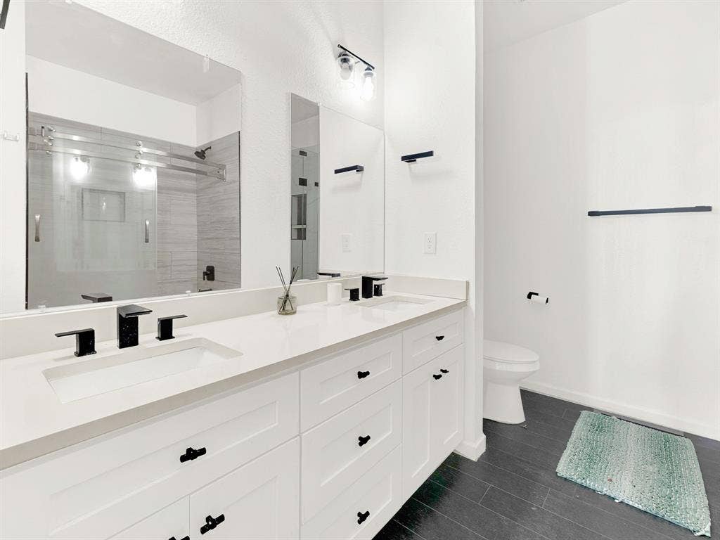 Dual-sink vanity with white cabinetry, light-toned stone-surface countertop, matte black hardware, and dark-finish flooring