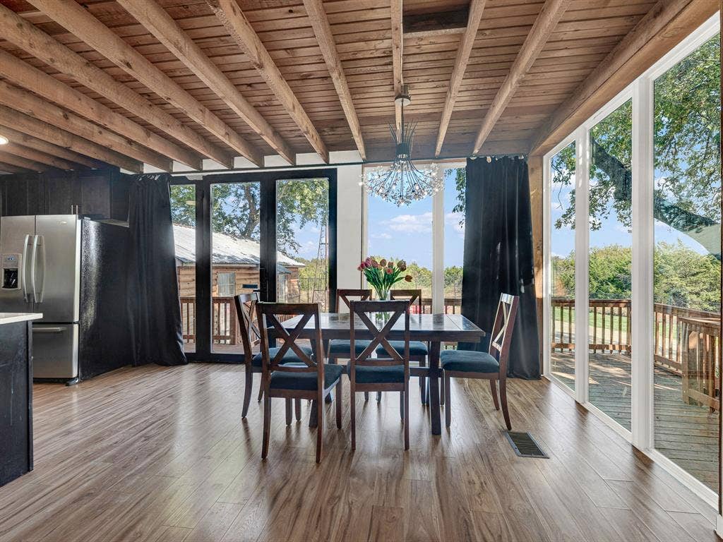 Exposed wood-beam ceiling, wood-finish flooring, floor-to-ceiling glass paneling, stainless steel refrigerator, and built-in modern chandelier lighting