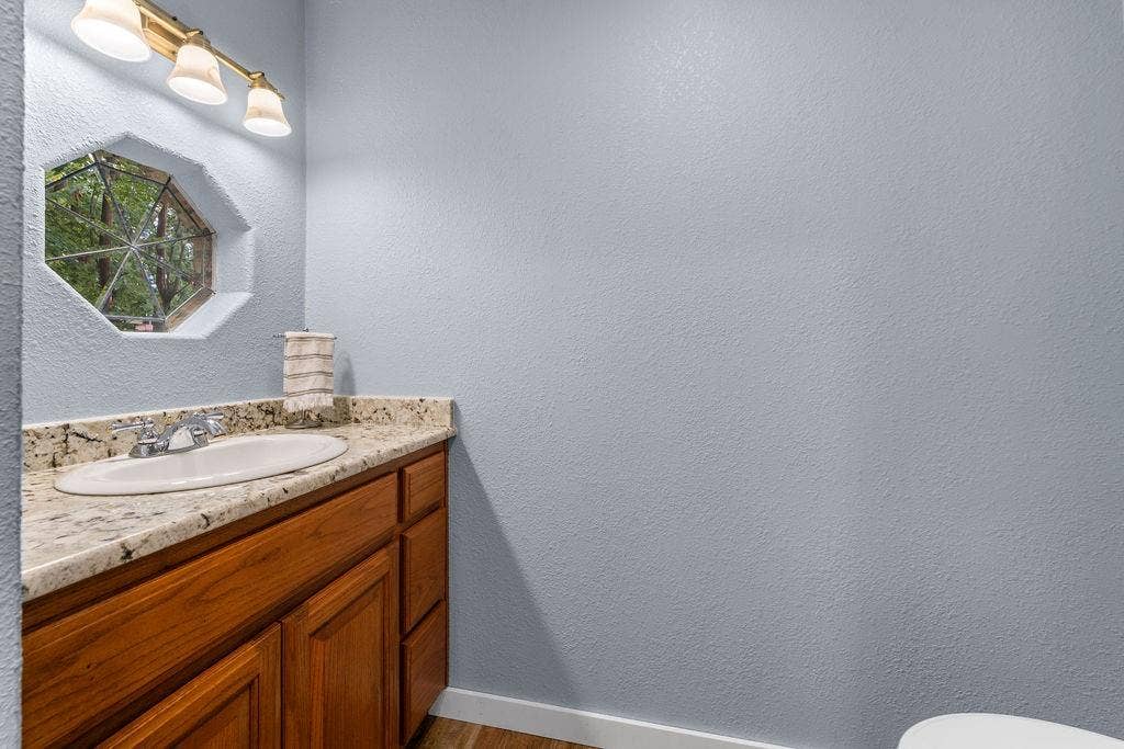 15993 Big Oak Bay Road, Tyler, TX 75707 Natural stone-surface vanity with an integrated oval sink, wood-finish cabinetry, hexagonal accent window, and three-bulb vanity lighting fixture