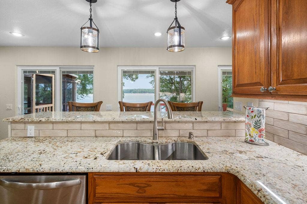 15993 Big Oak Bay Road, Tyler, TX 75707 Kitchen featuring stone countertops, tile backsplash, stainless steel dual-basin sink with gooseneck faucet, stained wood cabinetry, and pendant lighting