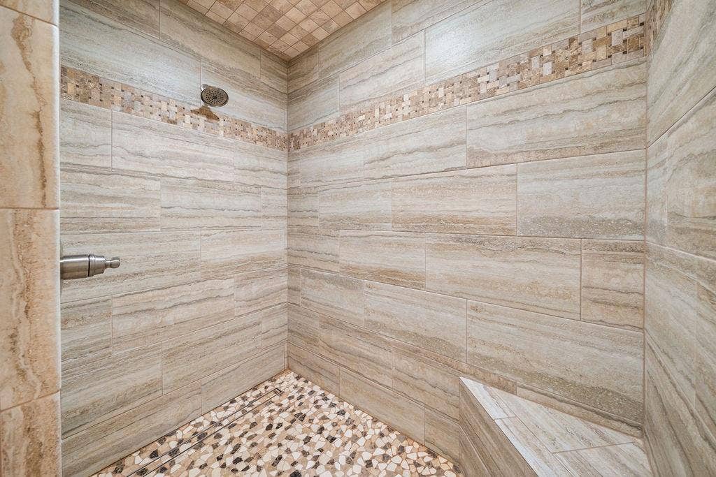 15993 Big Oak Bay Road, Tyler, TX 75707 Walk-in shower featuring floor-to-ceiling stone-finish wall tile, a decorative mosaic tile inlay, a built-in corner bench, a pebble-finish floor, and a metal shower head