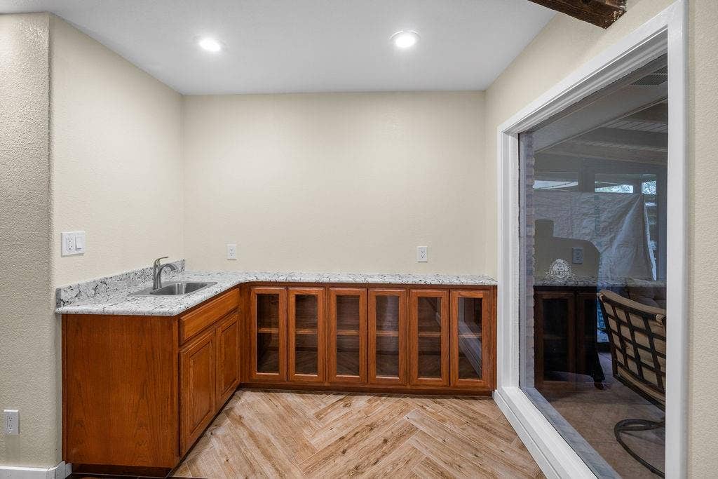 15993 Big Oak Bay Road, Tyler, TX 75707 Custom cabinetry featuring glass-panel doors, light-toned stone countertop, stainless steel undermount sink with arched faucet, and wood-finish flooring in a herringbone pattern