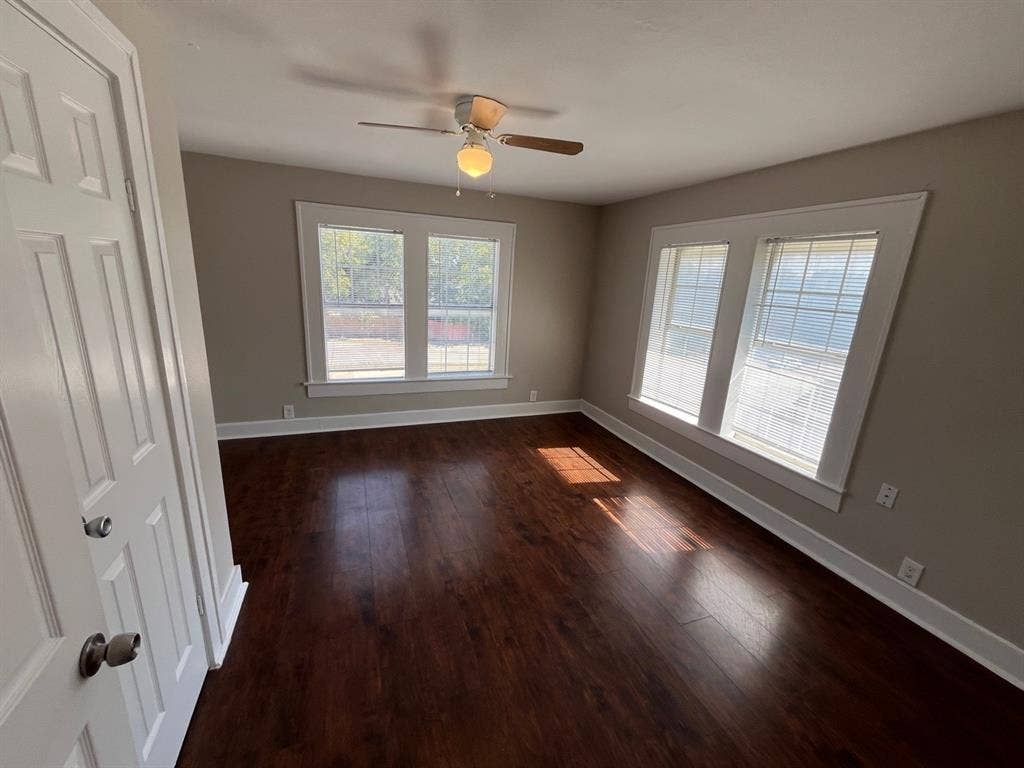 Interior space featuring dark wood-finish flooring, dual sets of windows with white trim, and a ceiling-mounted fan with integrated light fixture
