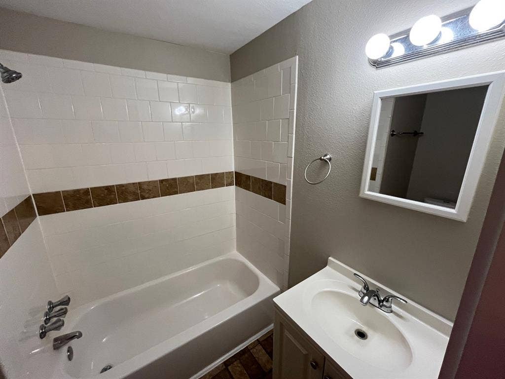 Bathroom featuring a tub-shower combination with decorative tile inlay, vanity with integrated basin, multi-bulb wall-mounted lighting, and a framed mirror