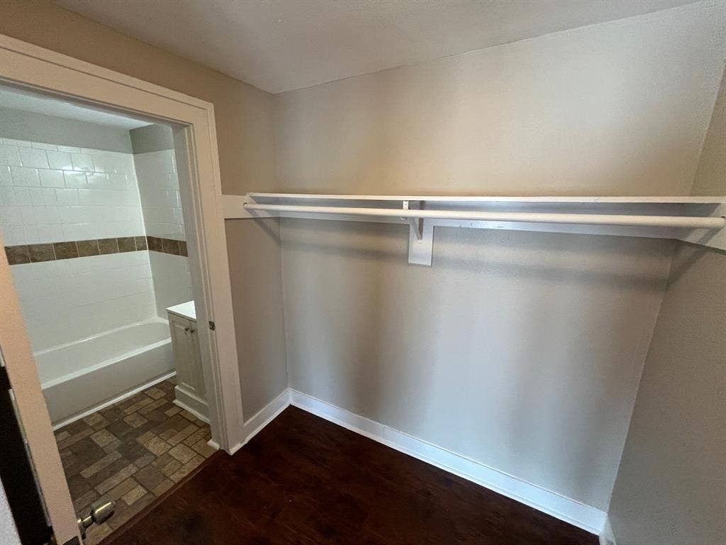 Storage area with wood-finish flooring and built-in shelving, featuring a doorway into a bathroom with a tile-surround tub and stone-tile flooring