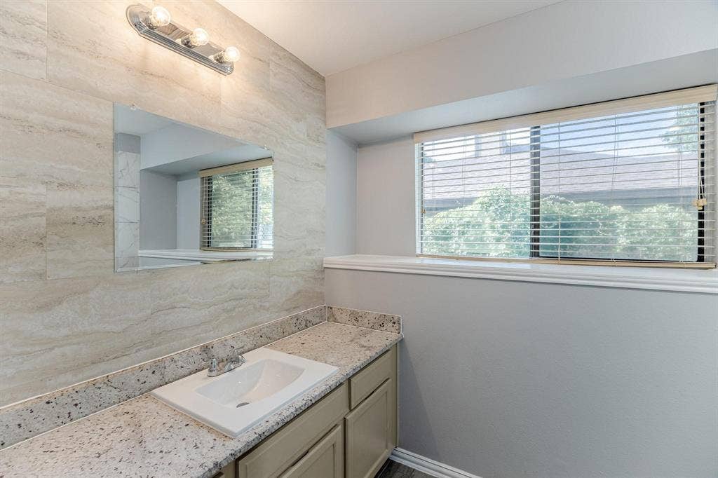 3618 Flagstone Avenue, Rowlett, TX 75088 Vanity featuring a stone-look countertop, integrated sink, neutral-toned cabinetry, floor-to-ceiling textured wall tile, and a three-bulb light fixture