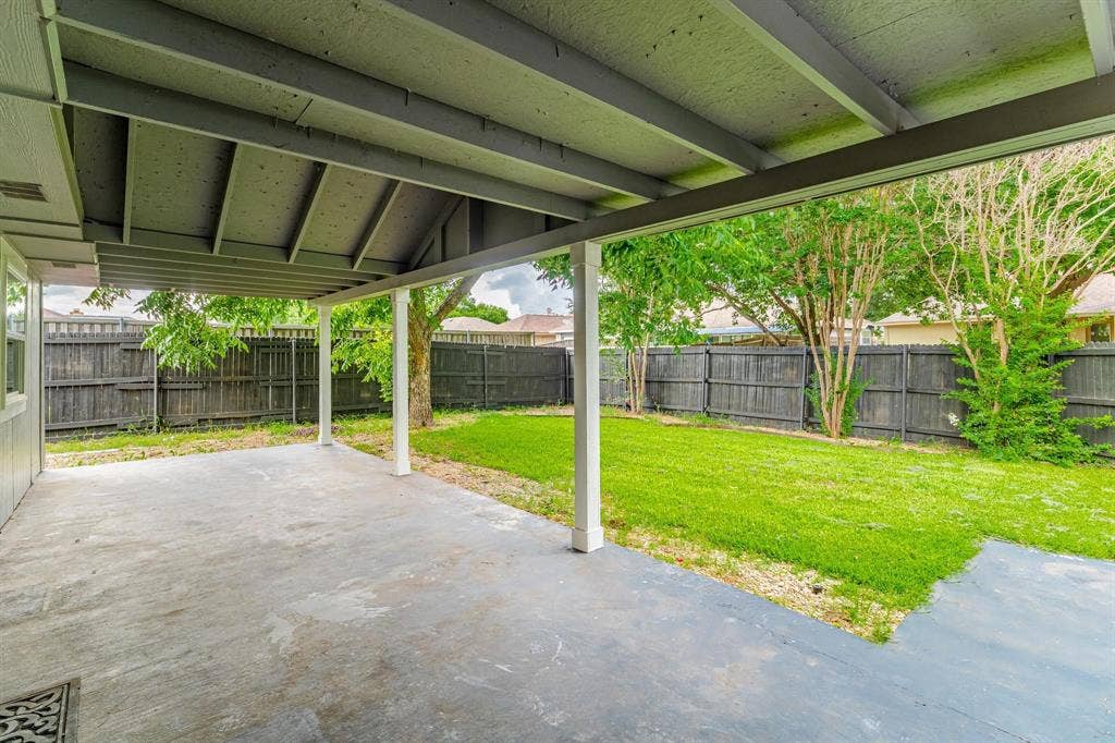 3618 Flagstone Avenue, Rowlett, TX 75088 Covered concrete patio featuring exposed rafters and square support columns, opening to a fenced backyard with a lawn and mature trees