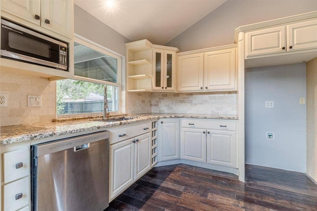 3618 Flagstone Avenue, Rowlett, TX 75088 Kitchen featuring cream-toned cabinetry with glass-front display sections, stone-finish countertops, subway-style backsplash, integrated stainless steel dishwasher, and wood-finish flooring