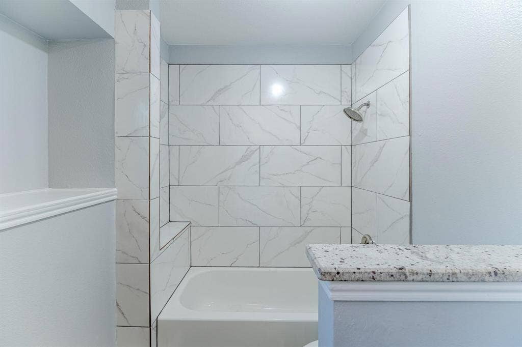 3618 Flagstone Avenue, Rowlett, TX 75088 Tub and shower combination featuring floor-to-ceiling large-format tile with veining, built-in recessed shelving, and a stone-topped half wall