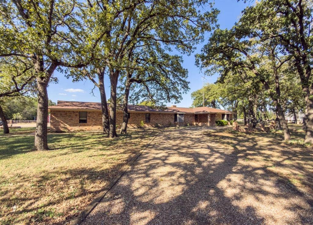 Single-story brick exterior residence featuring a sprawling gravel driveway and mature, expansive tree canopy