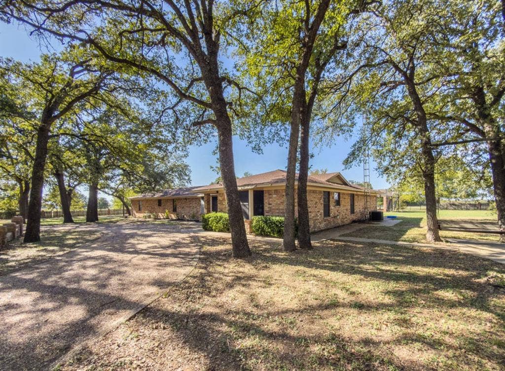 Single-story brick exterior featuring a sprawling lot, mature shade trees, and a gravel driveway