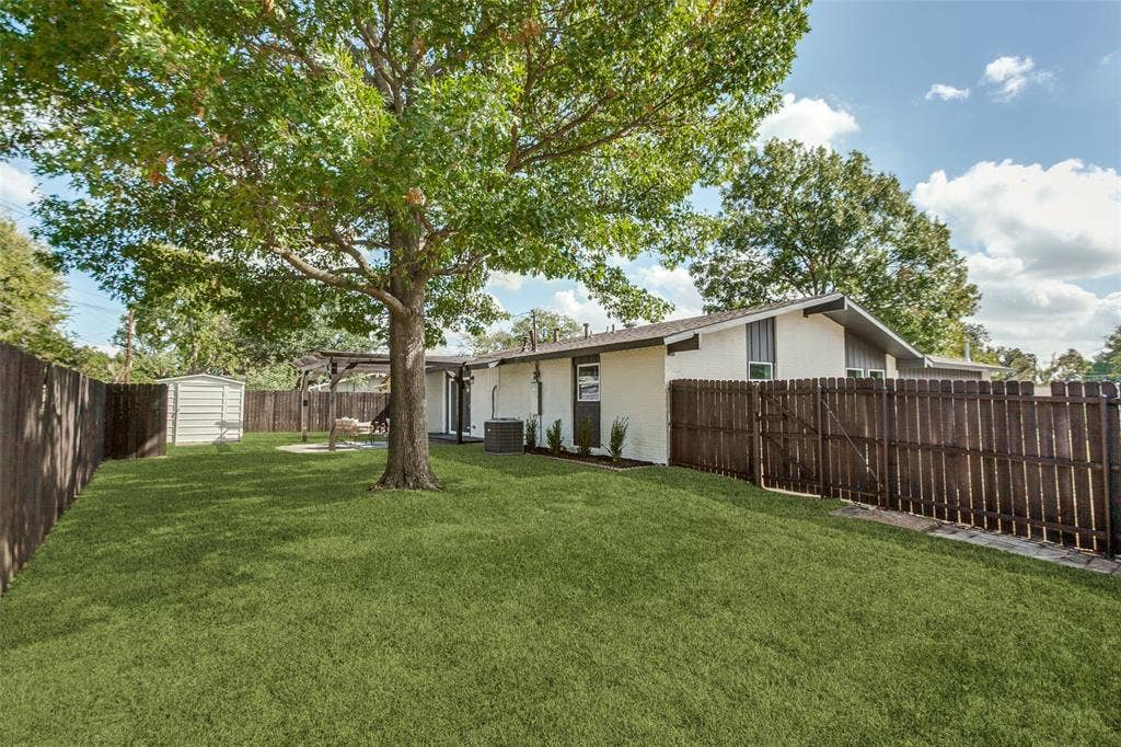 828 Melrose Drive, Richardson, TX 75234 Rear exterior featuring a spacious fenced yard, mature trees, a covered patio area, and a detached storage structure