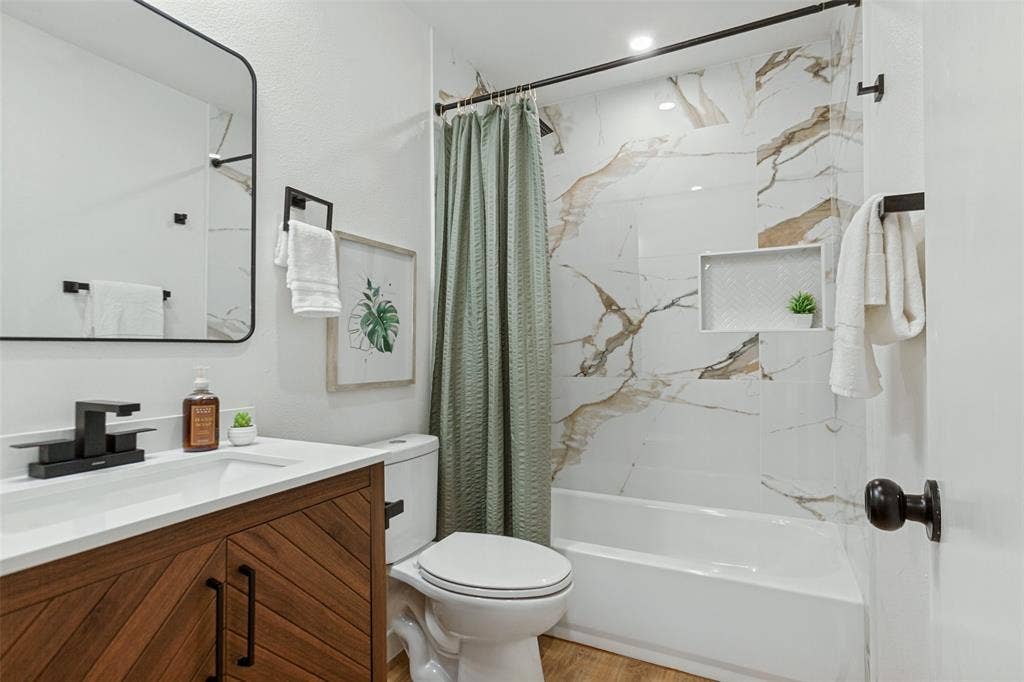828 Melrose Drive, Richardson, TX 75234 Bathroom featuring a wood-finish vanity with a rectangular sink, matte black faucet, wall-mounted mirror, and tub surround with decorative stone-patterned tile and a recessed niche
