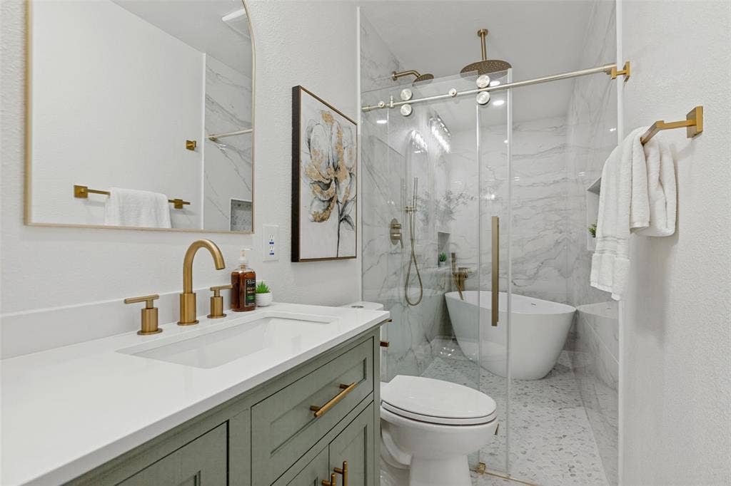 828 Melrose Drive, Richardson, TX 75234 Vanity with solid surface countertop, green cabinetry, brass hardware, arched wall mirror, and a walk-in wet room featuring stone-finish wall tile, a freestanding soaking tub, and a rainfall showerhead behind a sliding glass door