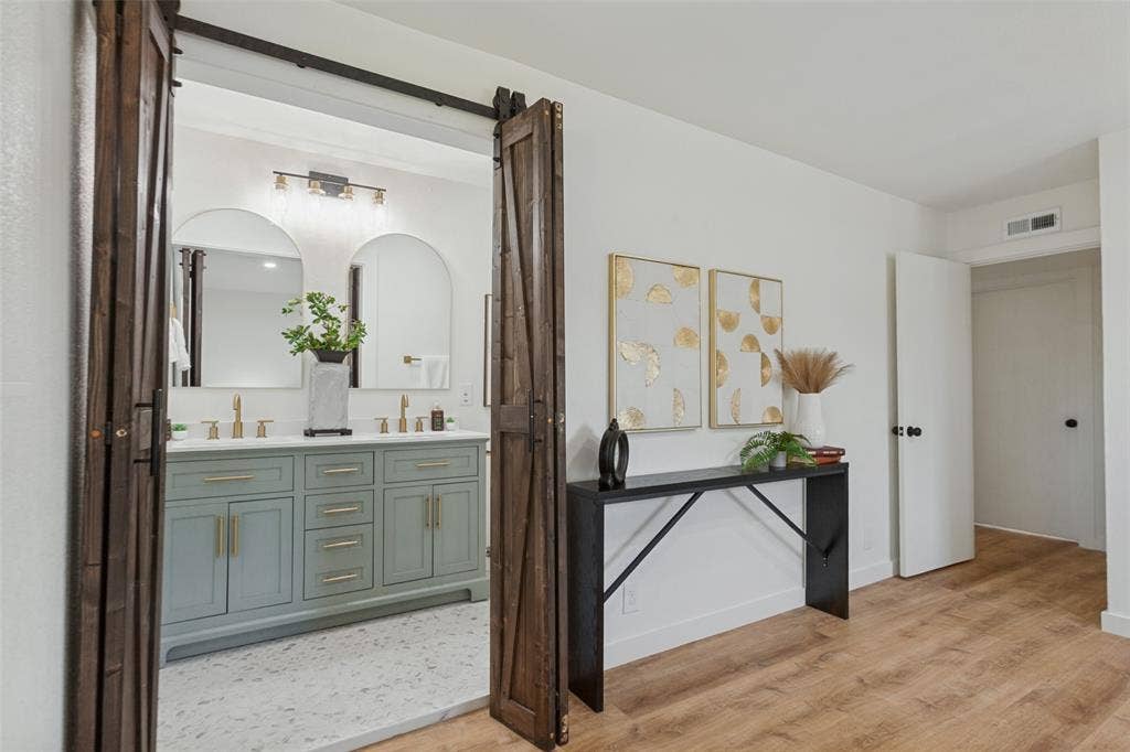 828 Melrose Drive, Richardson, TX 75234 Double vanity with a light-toned cabinet, brass hardware, arched mirrors, and wood-finish flooring, featuring a dark-stained sliding barn door entry