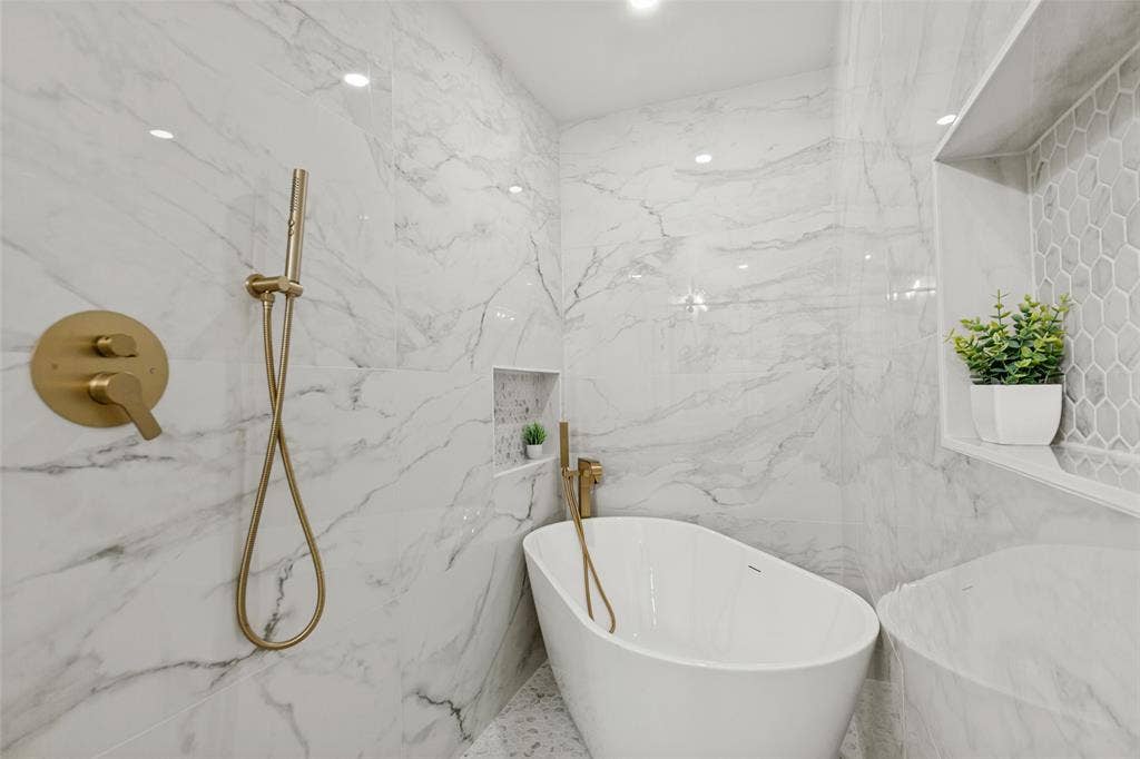 828 Melrose Drive, Richardson, TX 75234 Freestanding soaking tub featuring a metallic handheld faucet, wall-integrated shelving, and floor-to-ceiling large-format stone-finish tile surround