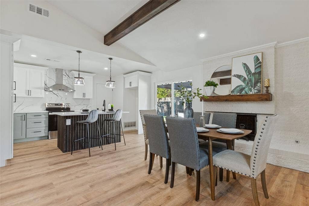 828 Melrose Drive, Richardson, TX 75234 Open-concept interior featuring vaulted ceilings with exposed wood beams, wood-finish flooring, kitchen island with vertical slat detailing, and a fireplace with a textured surround and heavy timber mantel