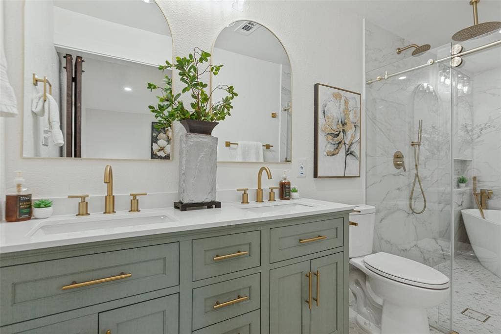 828 Melrose Drive, Richardson, TX 75234 Double vanity with green cabinetry and quartz countertop, dual arched wall mirrors, brass-finish fixtures, walk-in glass-enclosed shower, and freestanding soaking tub