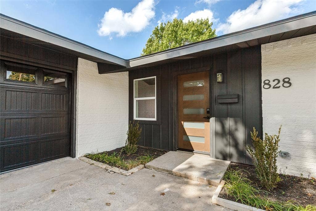 828 Melrose Drive, Richardson, TX 75234 Exterior entry featuring a contrast-painted brick and wood-panel facade, multi-panel garage door, modern wood-tone door with horizontal glass inserts, and concrete hardscaping