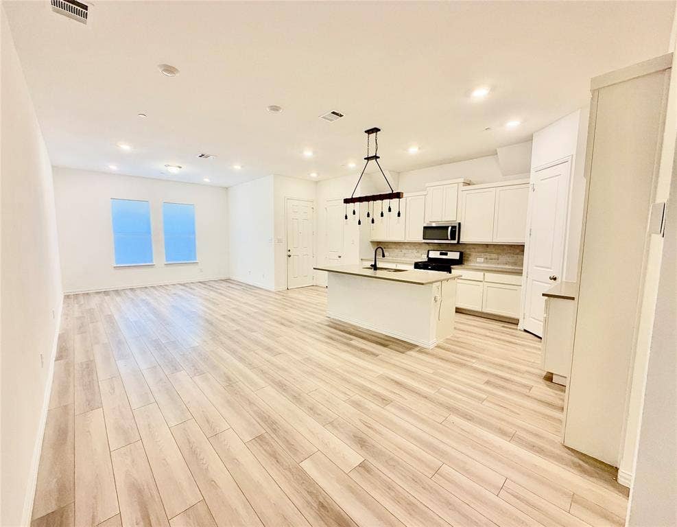 Open-concept kitchen and living area featuring wood-finish flooring, white shaker cabinetry, central island with an integrated sink, recessed lighting, and a linear pendant fixture