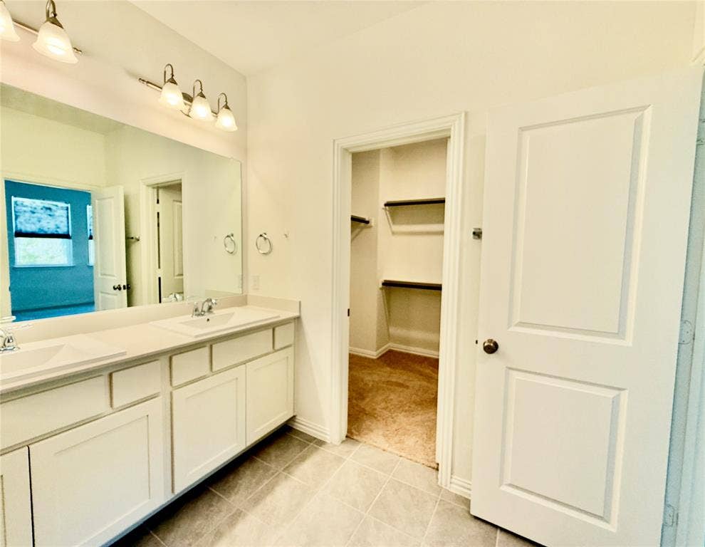 Dual-sink vanity with white cabinetry, integrated countertop, and oversized wall-mounted mirror, accented by dual three-light fixtures and tile flooring