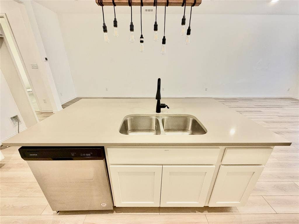 Kitchen island with light-toned solid surface countertop, stainless steel undermount double-basin sink, matte black gooseneck faucet, integrated stainless steel dishwasher, and suspended wood-beam pendant lighting with exposed filament bulbs over wood-finish flooring