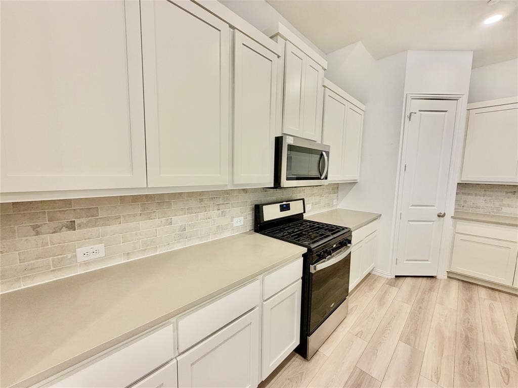 Kitchen featuring white shaker-style cabinetry, neutral-toned stone-finish countertops, subway tile backsplash, stainless steel microwave, and wood-finish flooring