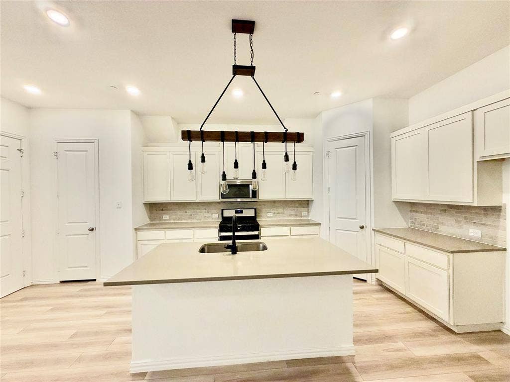 Kitchen featuring an island with a dual-basin sink, light-toned countertops, shaker-style cabinetry, a tile backsplash, and a multi-light wooden beam chandelier