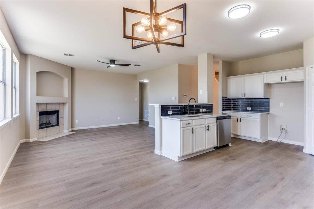 Open-concept interior featuring wood-finish flooring, a kitchen island with a dark tile backsplash, white cabinetry, a geometric statement chandelier, and a recessed fireplace with a neutral tile surround