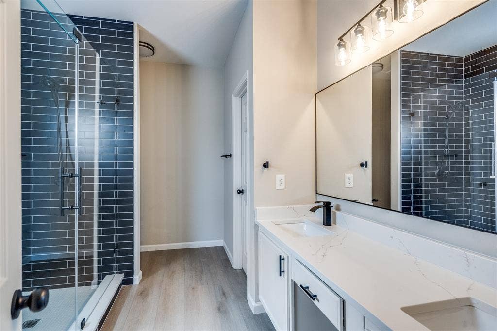 Bathroom featuring dual-sink vanity with light-toned countertop, walk-in shower with dark brick-style tile surround, glass enclosure, and wood-finish flooring