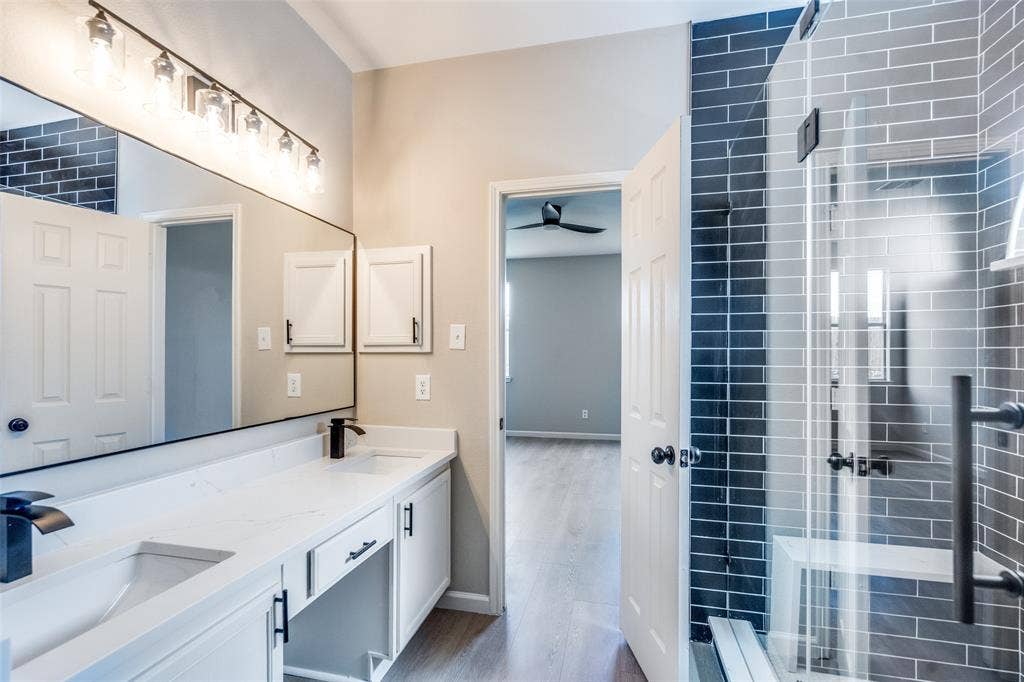 Dual-sink vanity with quartz countertop, walk-in shower featuring glass enclosure and dark ceramic wall tile, full-width vanity mirror, and matte black hardware