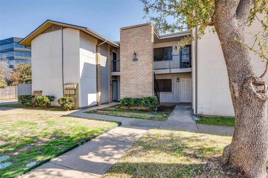 5623 Harvest Hill Road, #2023, Dallas, TX 75230 Exterior featuring light-colored smooth siding, brick facade accents, concrete walkways, and second-level metal balcony railings