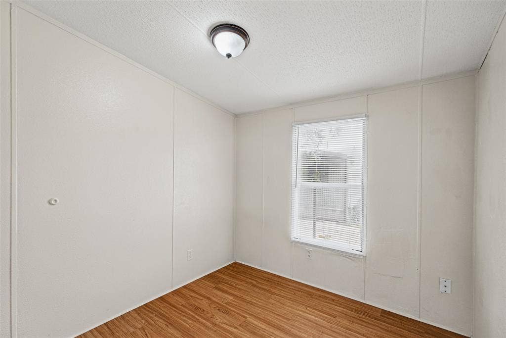 Interior room featuring wood-finish flooring, white panel walls, a single-window configuration with horizontal blinds, and a dome-style flush-mount ceiling fixture