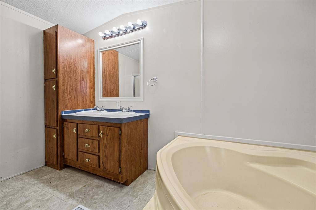 Bathroom featuring a dual-sink vanity with wood-finish cabinetry, a full-height storage cabinet, a framed mirror, and a soaking tub