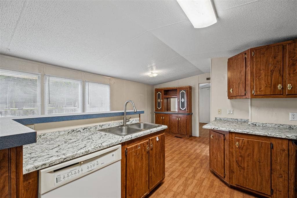 Kitchen featuring wood-finish cabinetry, stone-pattern laminate countertops, a dual-basin stainless steel sink with a high-arch faucet, built-in dishwasher, and wood-finish flooring