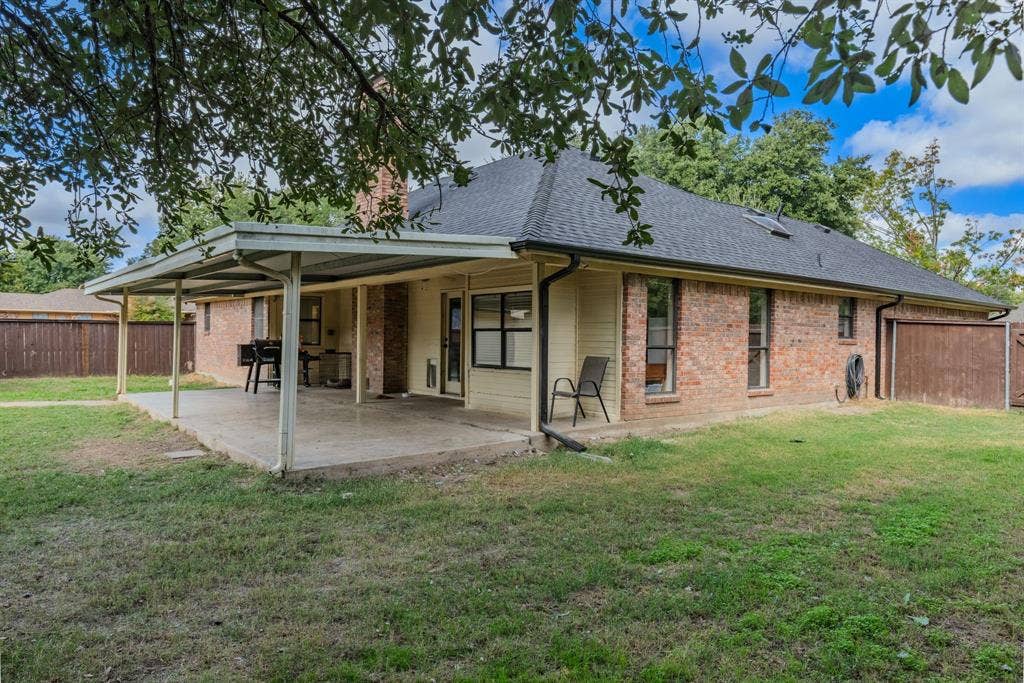 1307 Edna Street, Commerce, TX 75428 Rear exterior featuring a brick and siding facade, a covered patio, a chimney, a shingled roof, and a fenced backyard
